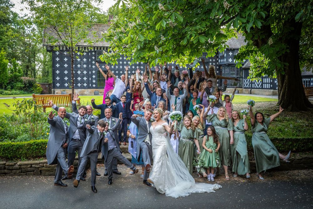 Bride and groom celebrate with large wedding party group photo outside Tudor venue.