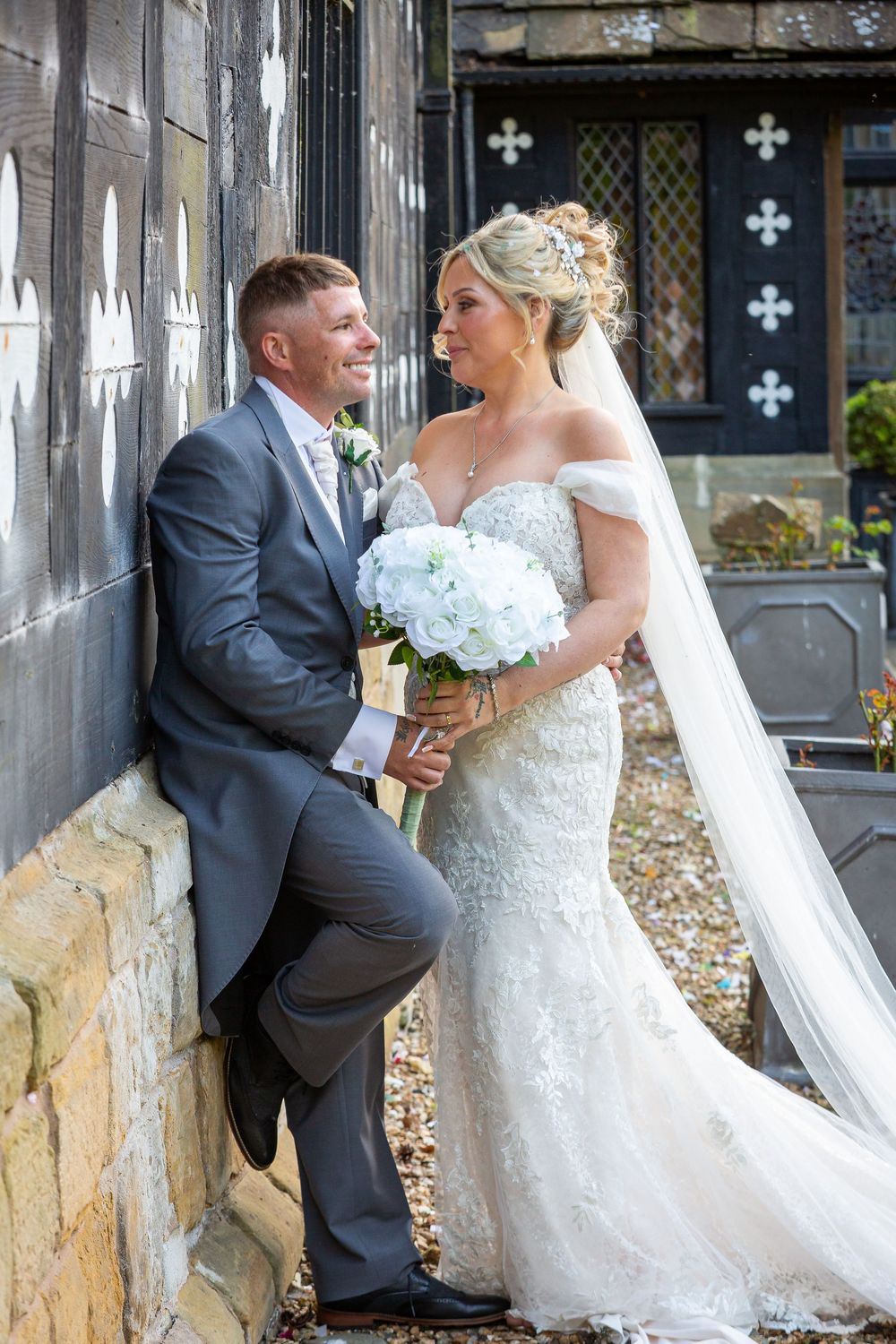 Bride in lace mermaid gown and groom in grey suit pose against historic stone church wall.