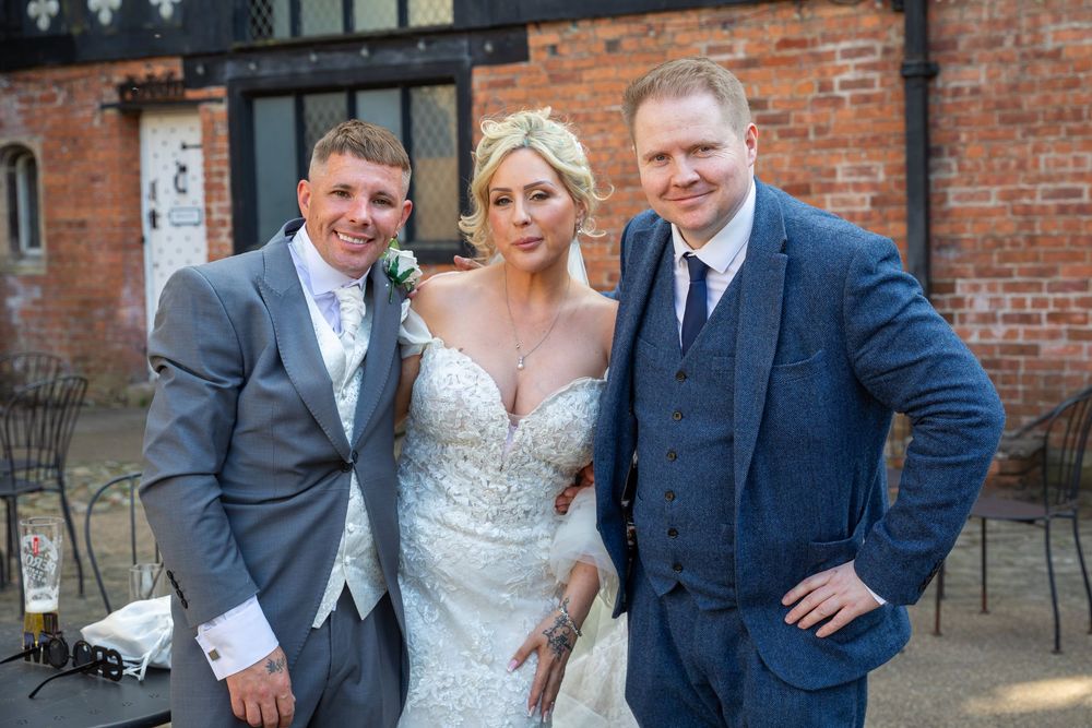 Bride in beaded gown poses with groom and guest outside rustic brick wedding venue.