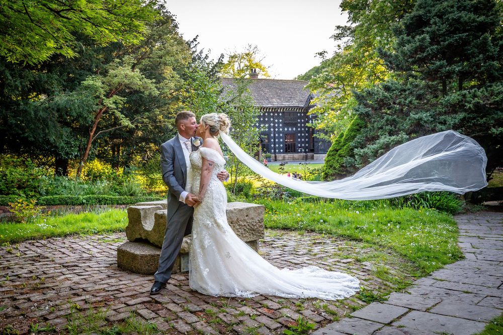 Bride and groom share a romantic kiss in a garden courtyard with the bride's veil flowing dramatically in the wind.