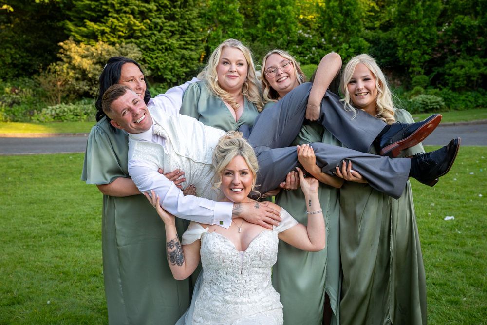 Bride laughs as bridesmaids in sage green dresses playfully lift groom on the lawn.