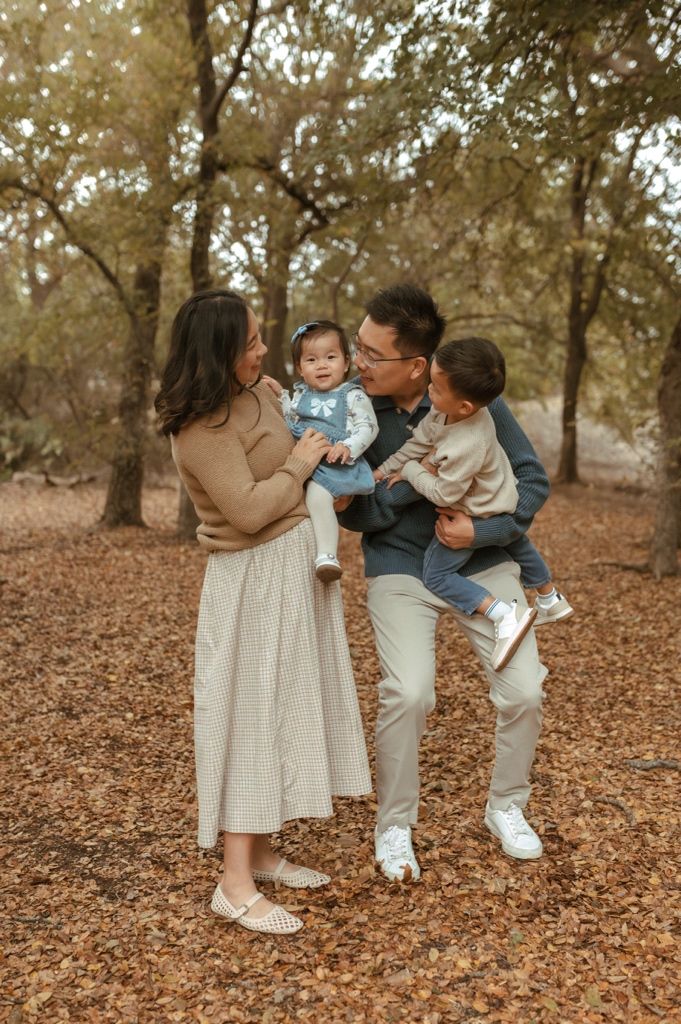 Family shares tender moments together while standing on fallen autumn leaves in a wooded park setting.