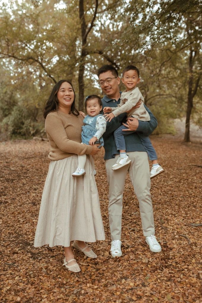 A family in coordinated beige and denim outfits poses together on fallen autumn leaves in a wooded setting.