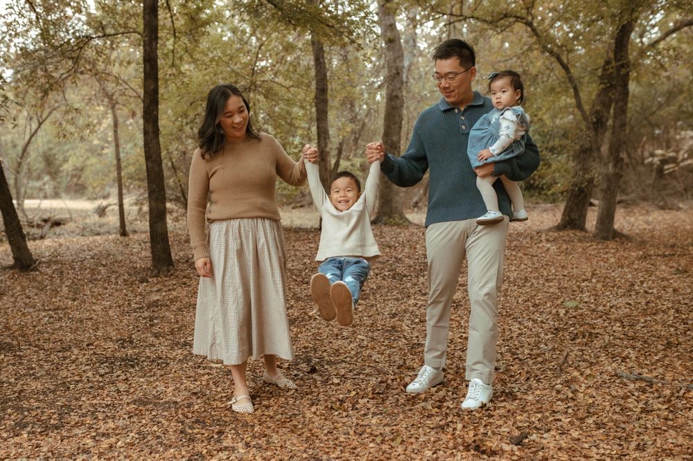 A family walks together through a forest path covered in autumn leaves on a sunny afternoon.