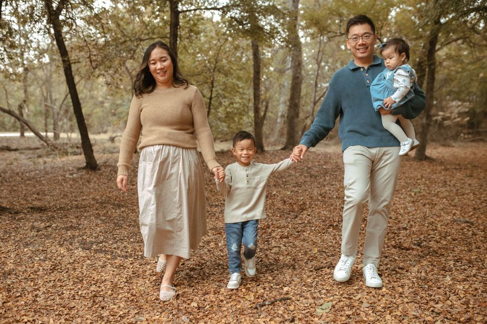 A family in coordinated beige and blue outfits walks together through a forest covered in fallen autumn leaves.