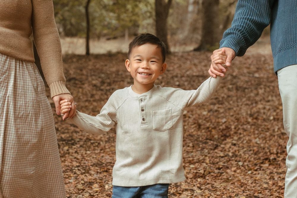 A child holds hands with parents while smiling in a forest setting with fallen leaves.