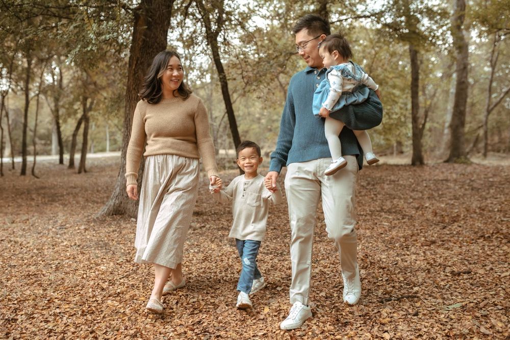 A family takes an autumn stroll together through a wooded park covered in fallen leaves.