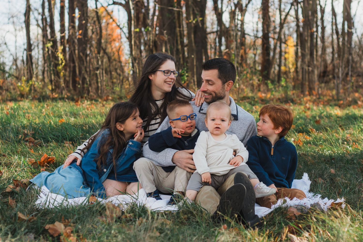Happy family of five sits together on a blanket in an autumn park, parents with three young children.