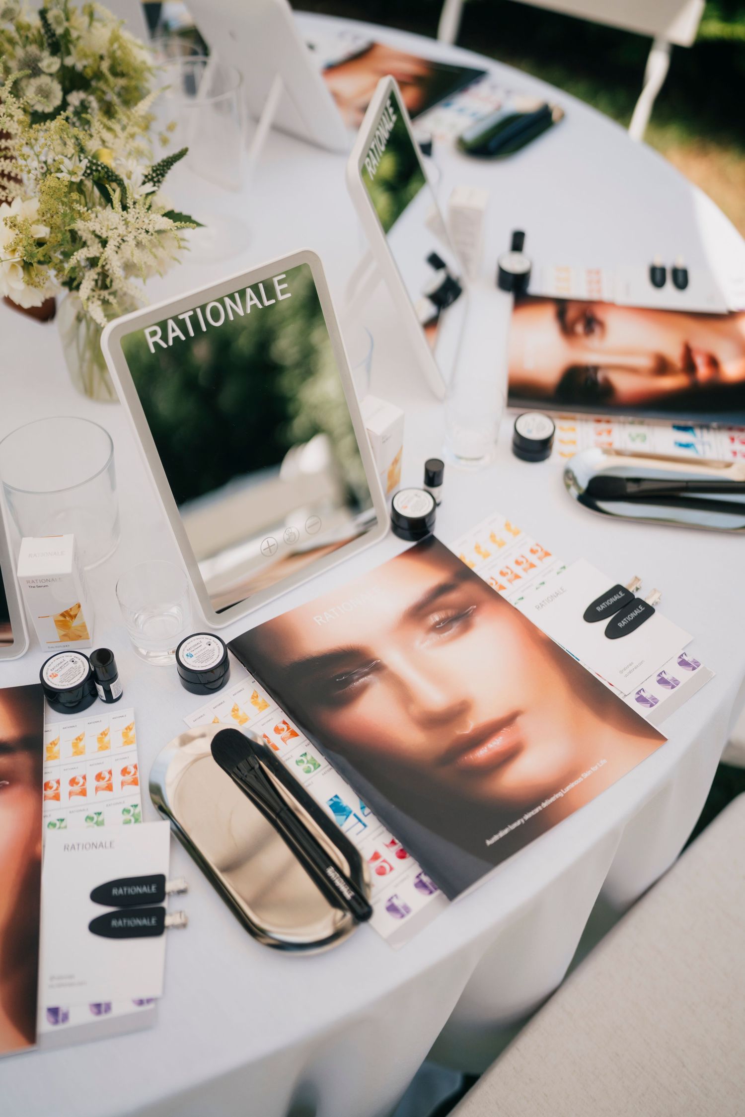 Makeup products and promotional photos scattered on a white surface with cosmetic bottles and beauty accessories.