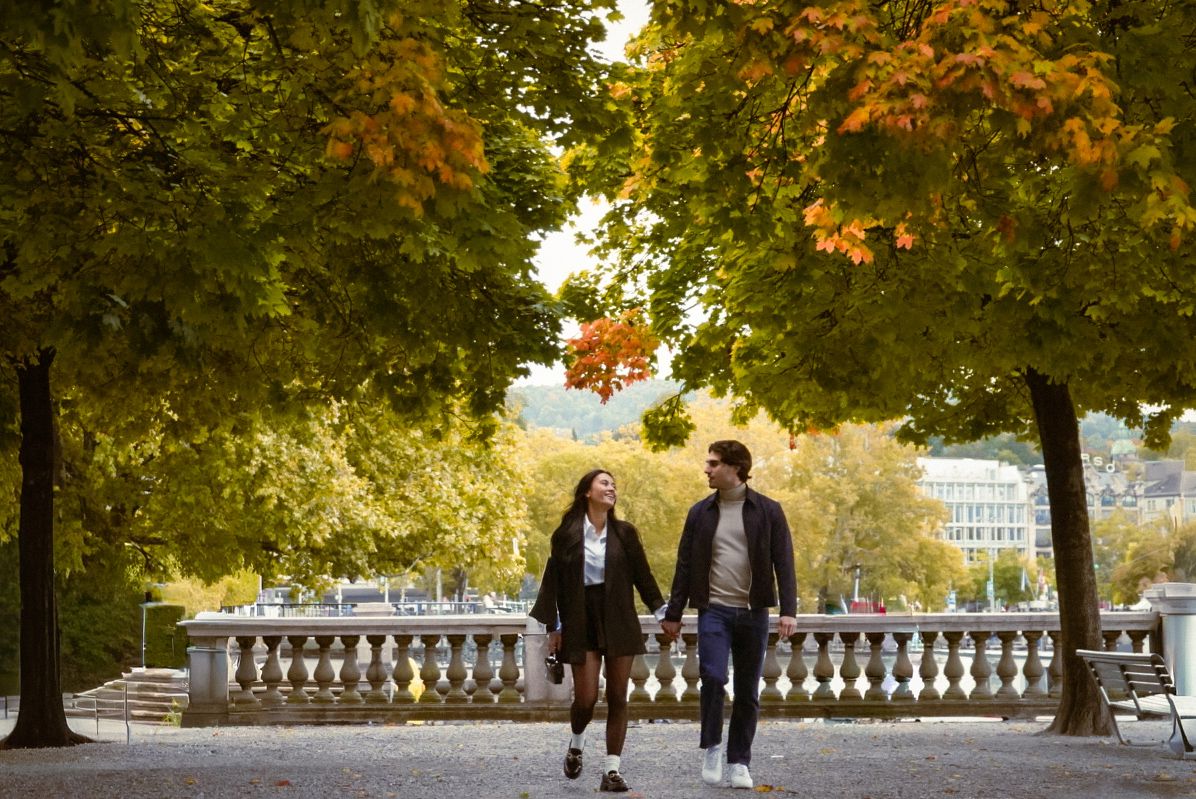A couple walks together on a stone balustrade path surrounded by autumn trees with golden leaves.
