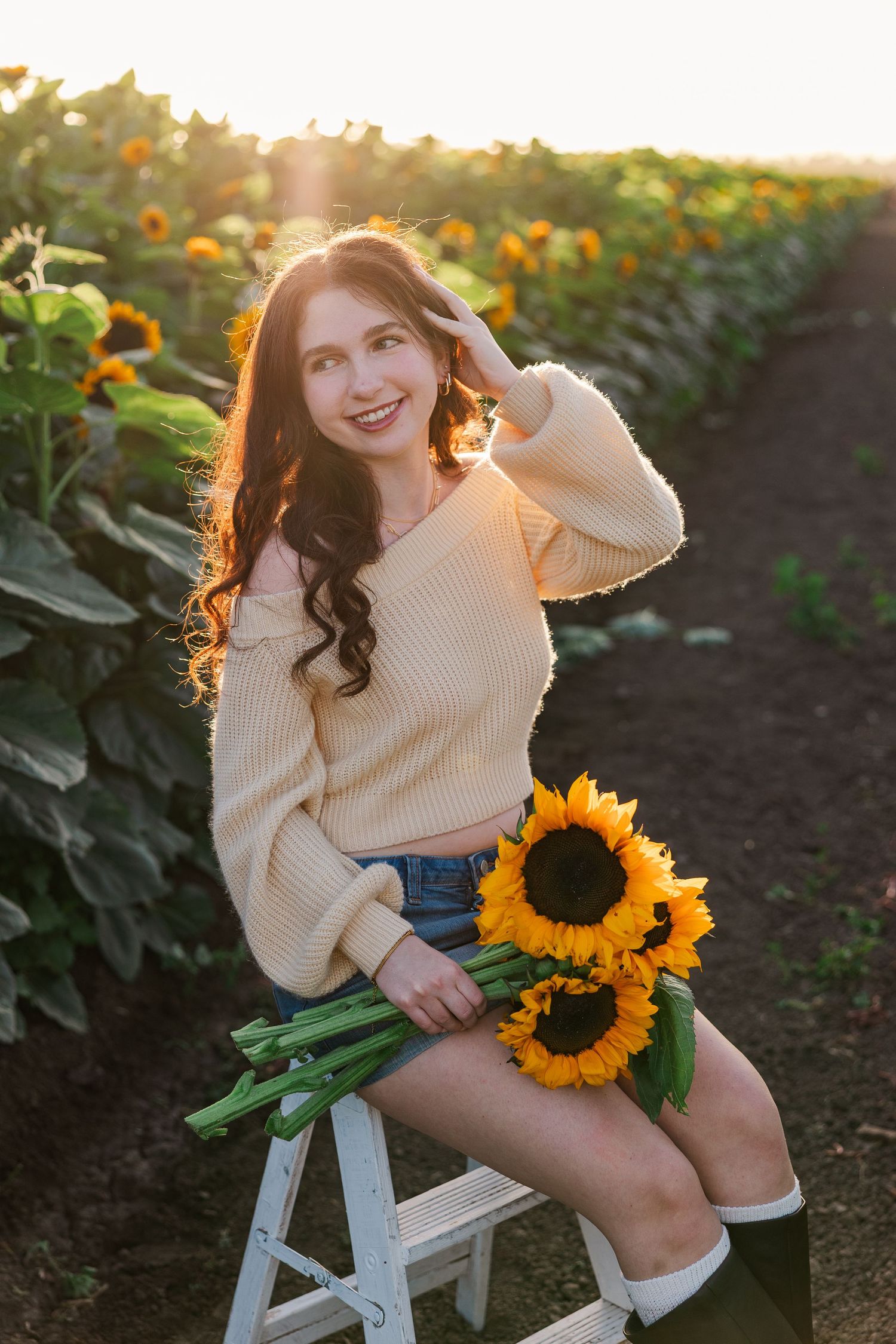 A person in a cream sweater sits on a white stool holding sunflowers during golden hour in a garden.