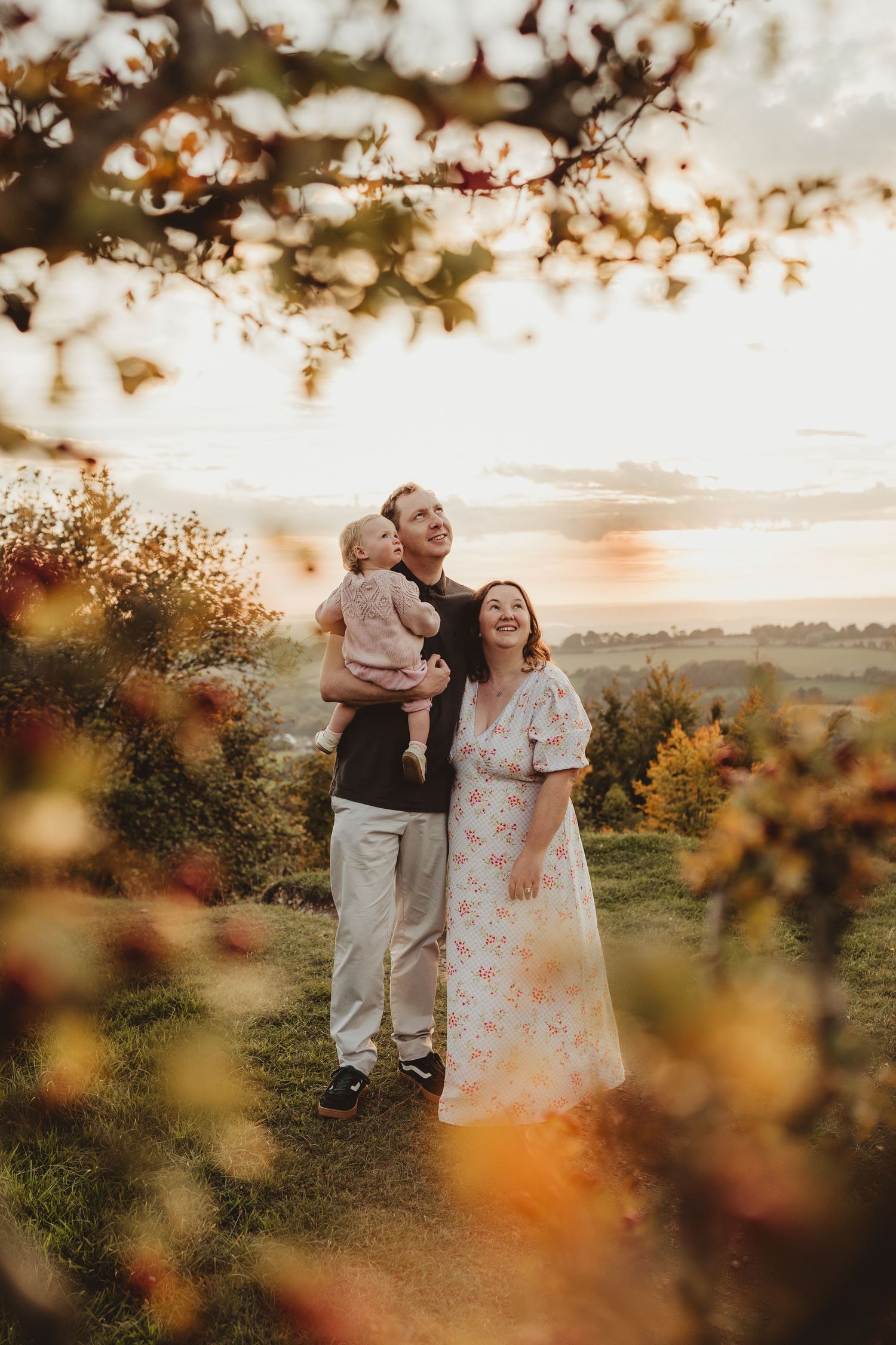A family enjoys a golden sunset moment together on a hilltop surrounded by autumn foliage and bokeh lighting effects.