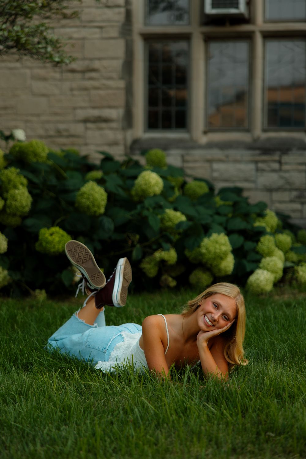 A person in casual attire lies on lush green grass near a stone building with hydrangea bushes in bloom.