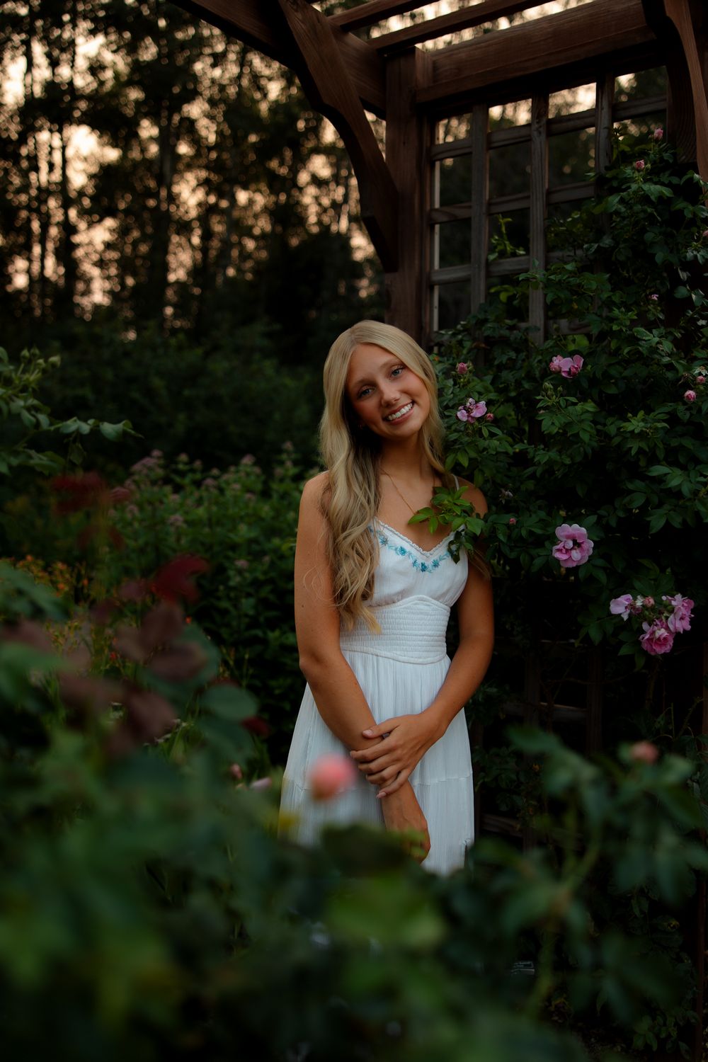 A white summer dress stands out against a garden backdrop with pink flowers during golden hour.