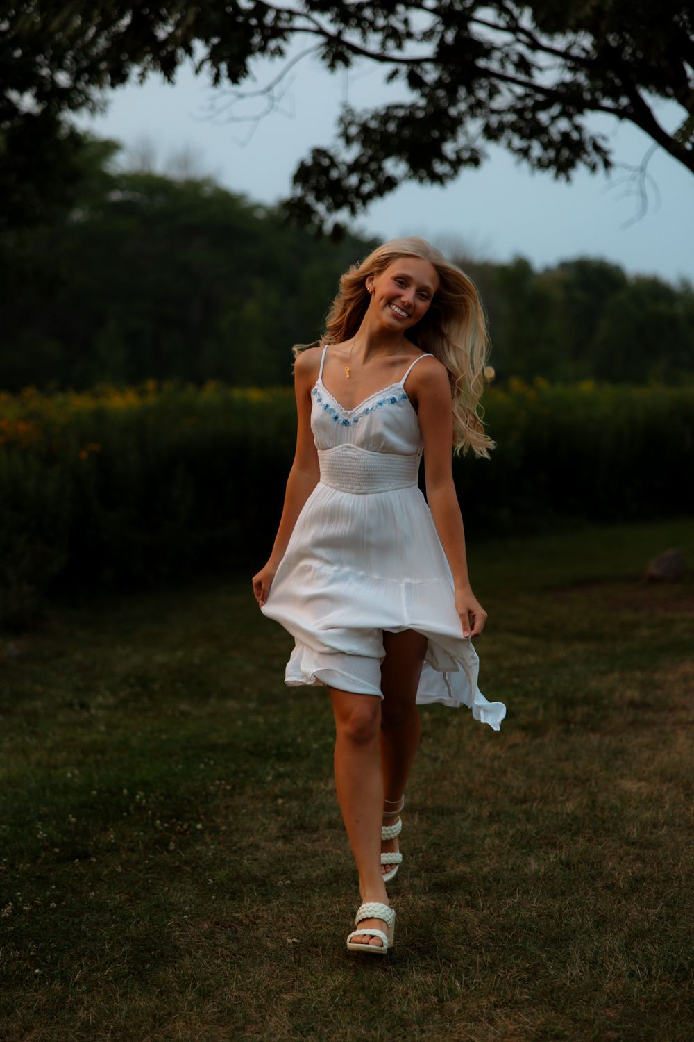 A woman in a flowing white sundress dances joyfully in a garden setting at dusk.