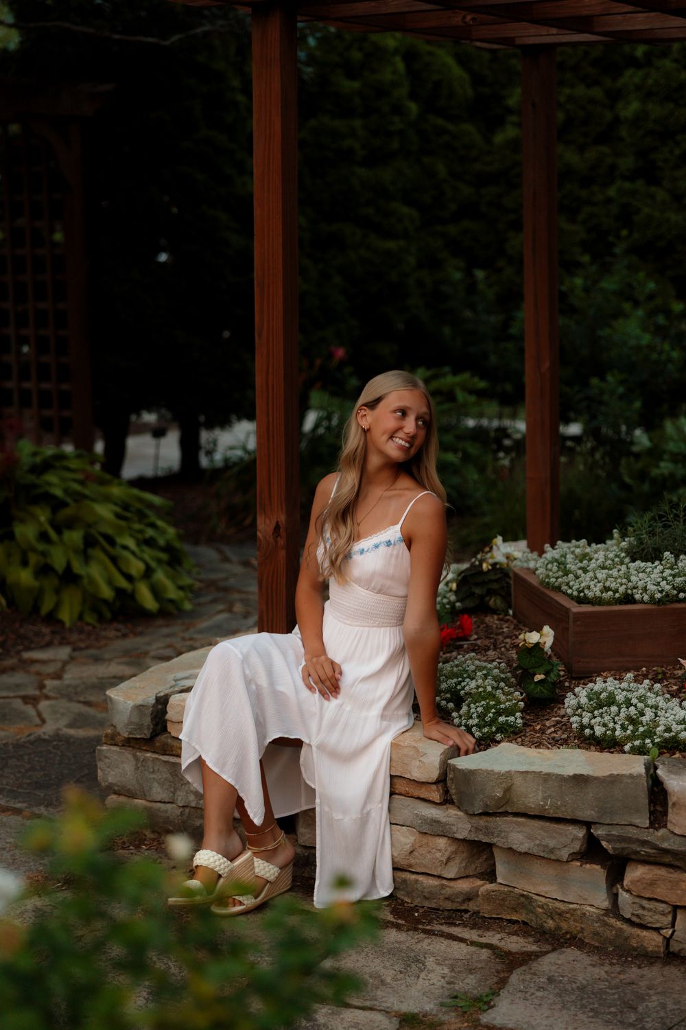 A woman in a white summer dress sits on stone steps near a wooden pergola surrounded by flowers.