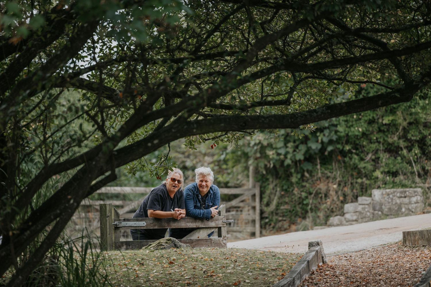Two elderly people relax at a wooden picnic table beneath lush green trees in a peaceful garden setting.
