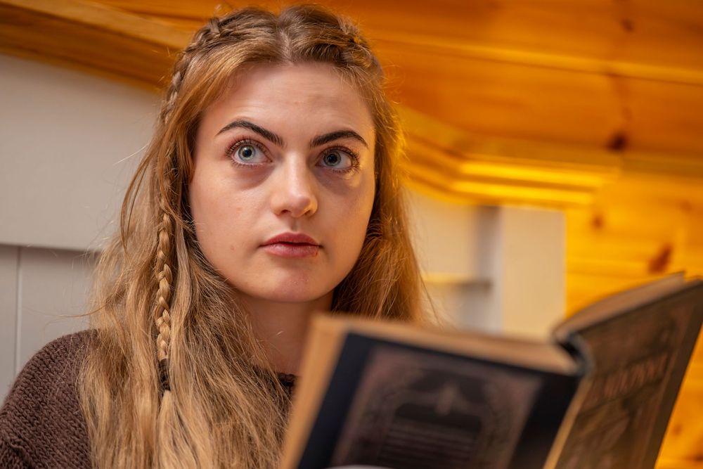 Young woman with braided blonde hair reading a book, looking up with wide blue eyes in a cozy wooden interior.