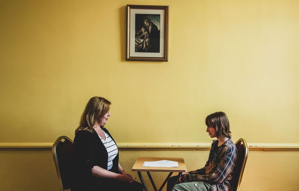Two people sit across from each other at a small round table against a yellow wall with a framed photo.