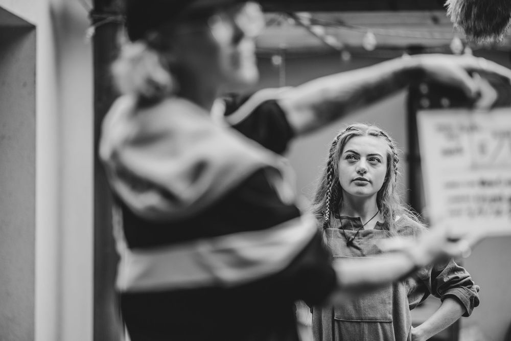 Black and white photo of a young woman watching dancers rehearse in a studio.
