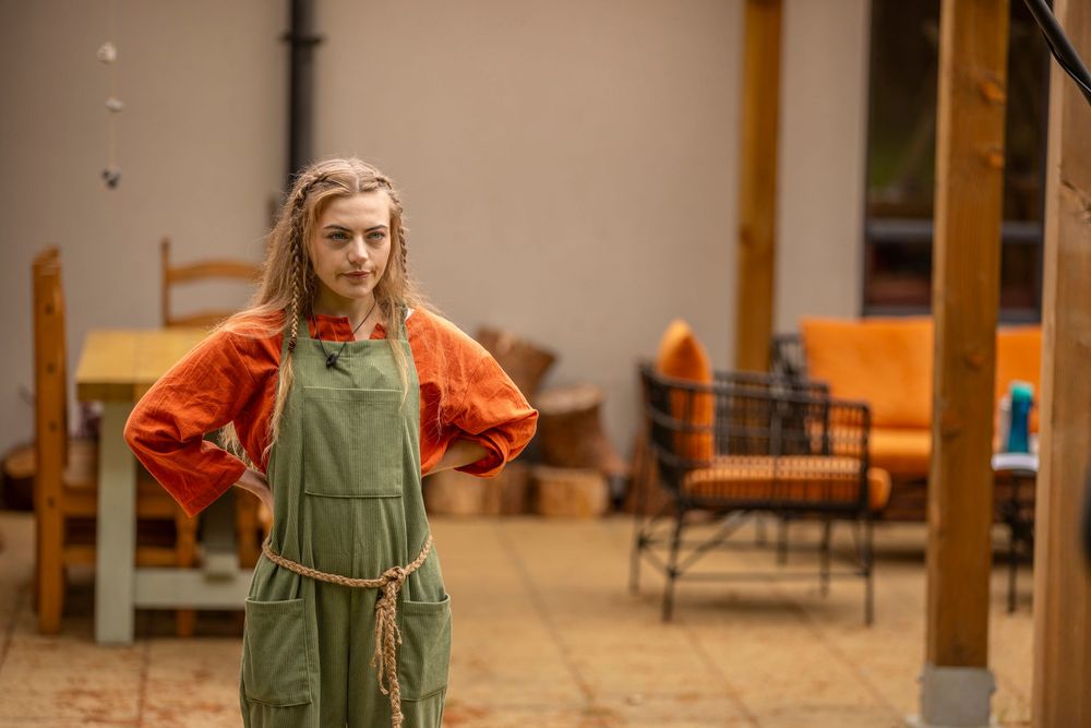 Young girl with braids wearing green overalls and orange blouse stands confidently on a patio.