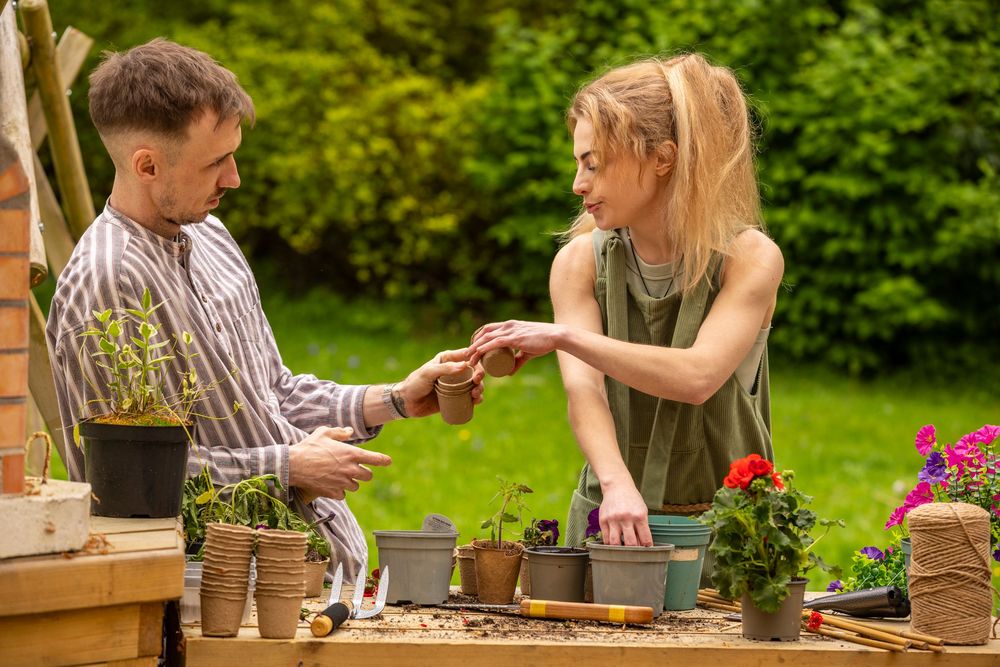 A young couple potting plants together at an outdoor garden table surrounded by flowers and greenery.