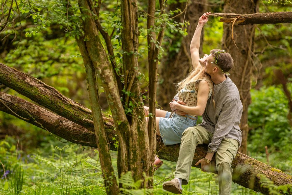 A couple sits romantically on a fallen log in a lush green forest, sharing a tender kiss outdoors.