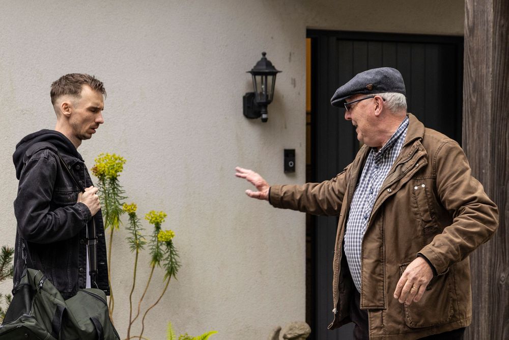 Two men having a conversation outside a house door, one gesturing with an outstretched hand.
