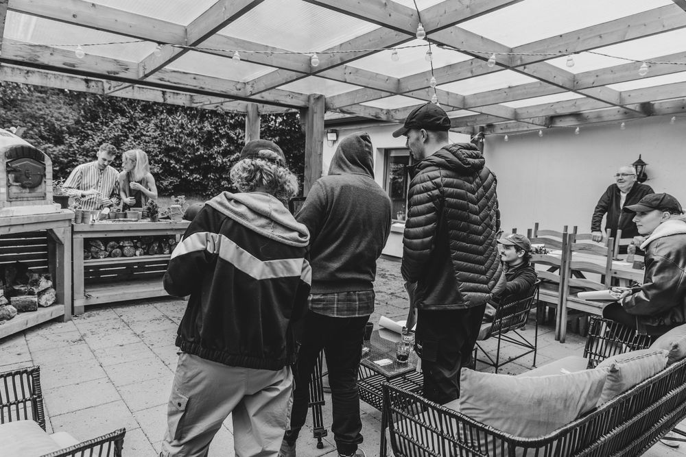 Black and white photo of a group of young people gathered outdoors under a pergola at a casual social event.