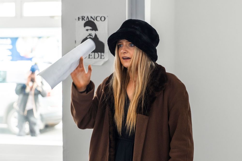 Person in brown coat and black hat walking outdoors while holding a beverage cup.