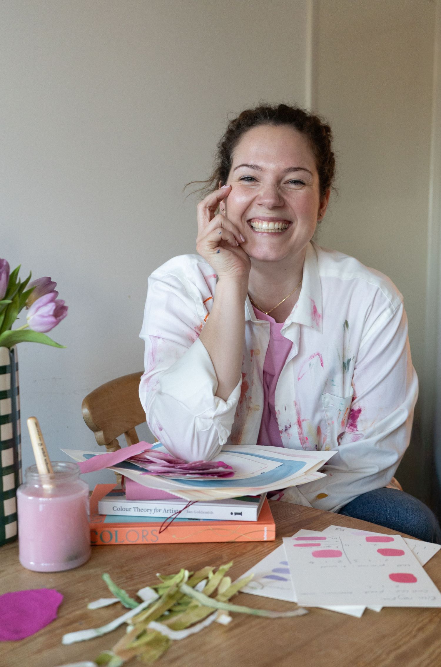 Smiling female artist with curly hair holding colourful fabric rolls, wearing a striped pink shirt and jeans. Branding portrait photoshoot.
