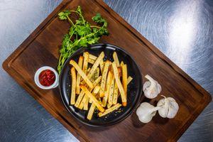 Fresh french fries with parsley garnish in a black dish on wooden serving board with garlic cloves.