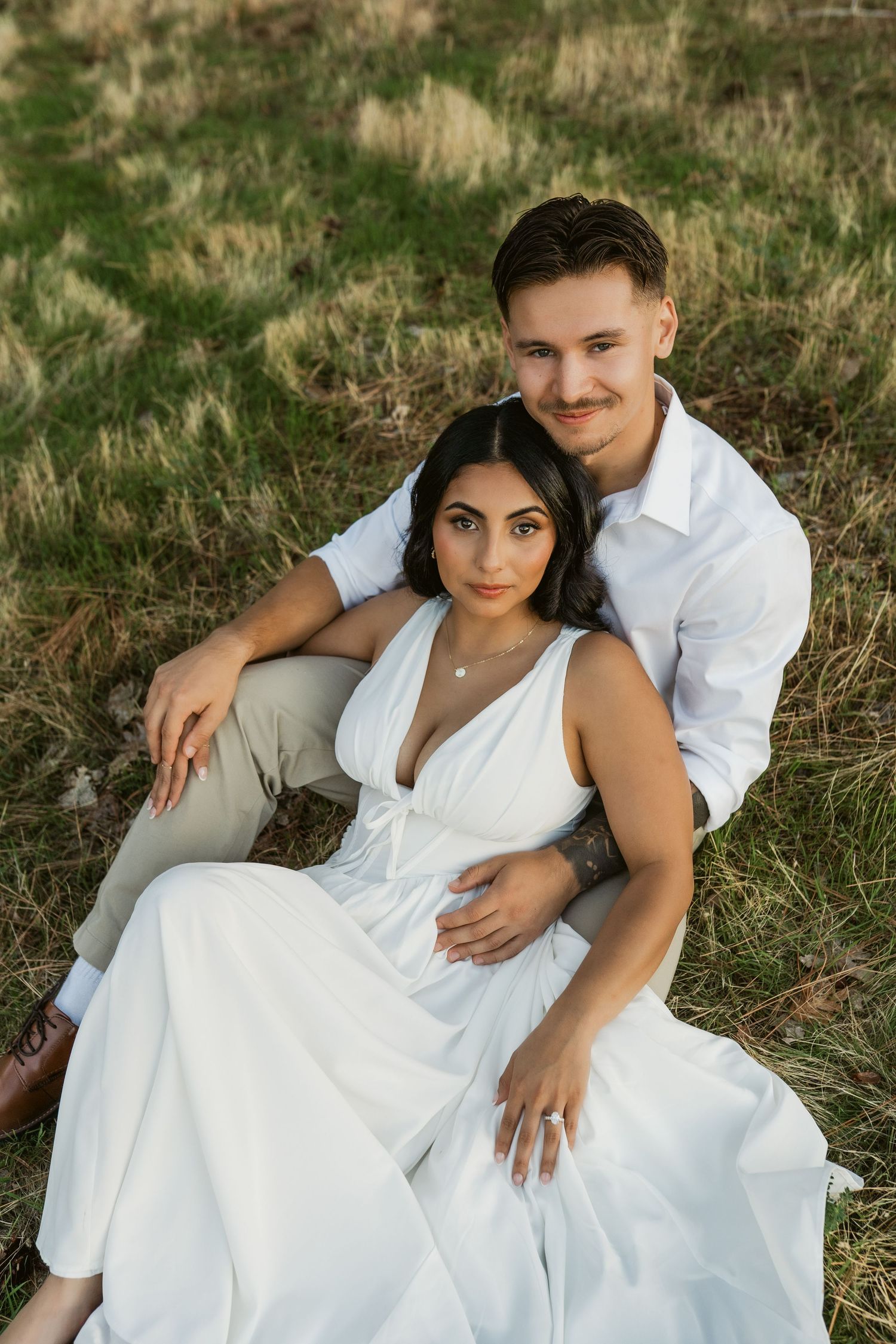 A romantic couple in white clothing poses together on grassy ground during a photo session.