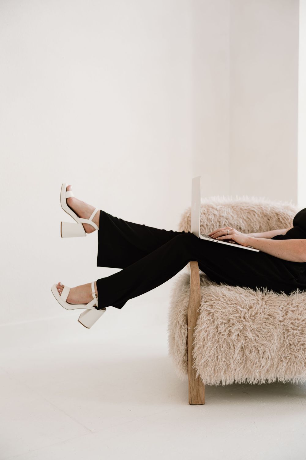 Person lounging on fluffy chair wearing black pants and white block heel sandals in minimalist setting.