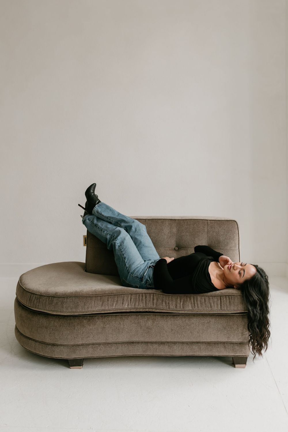 Person lying on a taupe velvet chaise lounge wearing jeans and black top against white wall in minimalist setting.