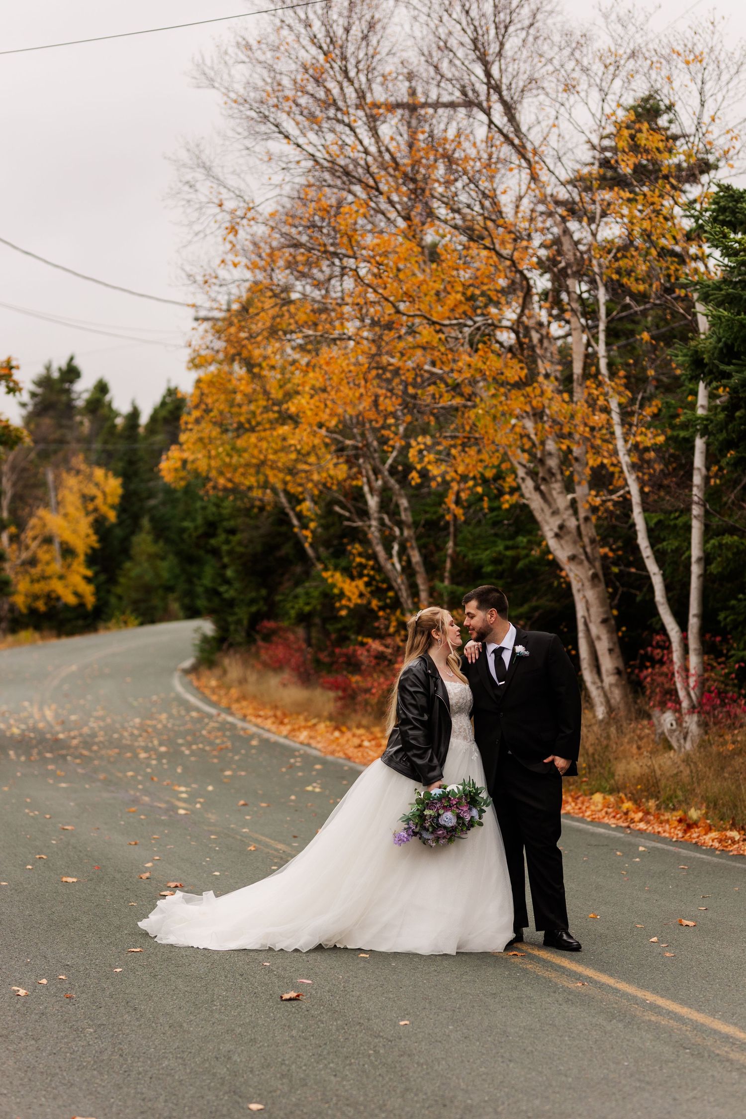 A couple shares a romantic moment on a winding autumn road lined with vibrant yellow and orange trees.