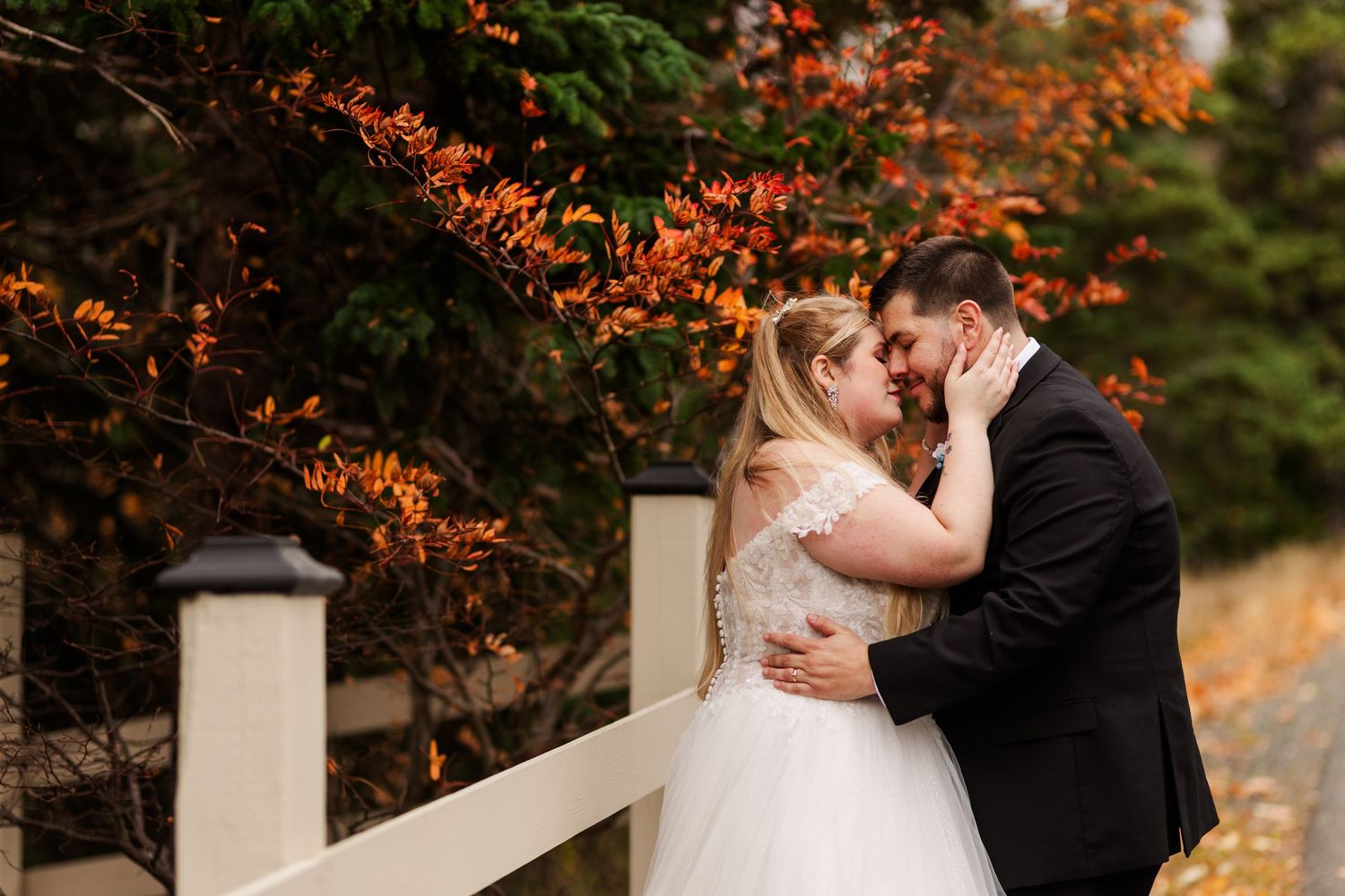 A romantic wedding couple embraces under vibrant autumn foliage near a white fence during an outdoor ceremony.