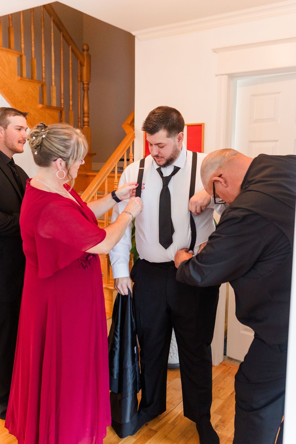 A group helps adjust a black tie and suspenders on a person in formal attire near a wooden staircase.