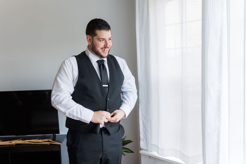 Professional in a dark vest and white shirt smiles while standing by bright windows in an elegant setting.