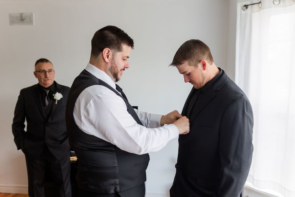 Groomsmen help each other get ready before a wedding ceremony in a bright white room.