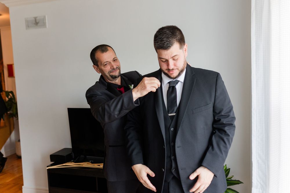 Two people in suits share a lighthearted moment while getting ready, with one adjusting the other's suit jacket.