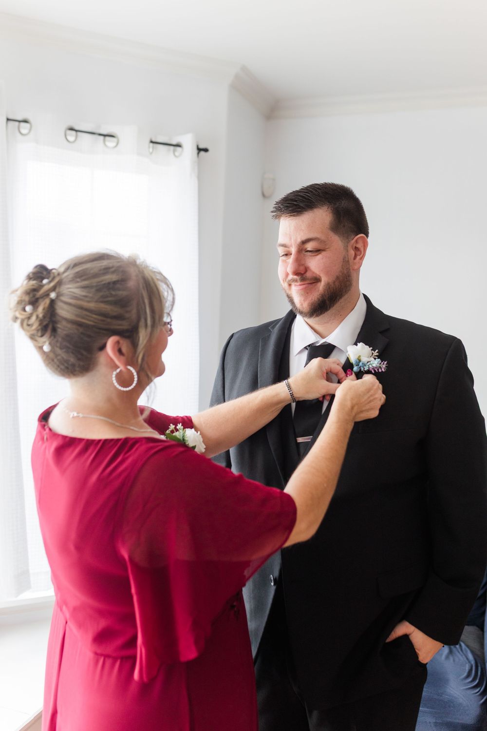 A person in a red dress helps adjust a boutonniere on a groom's suit jacket during wedding preparations.