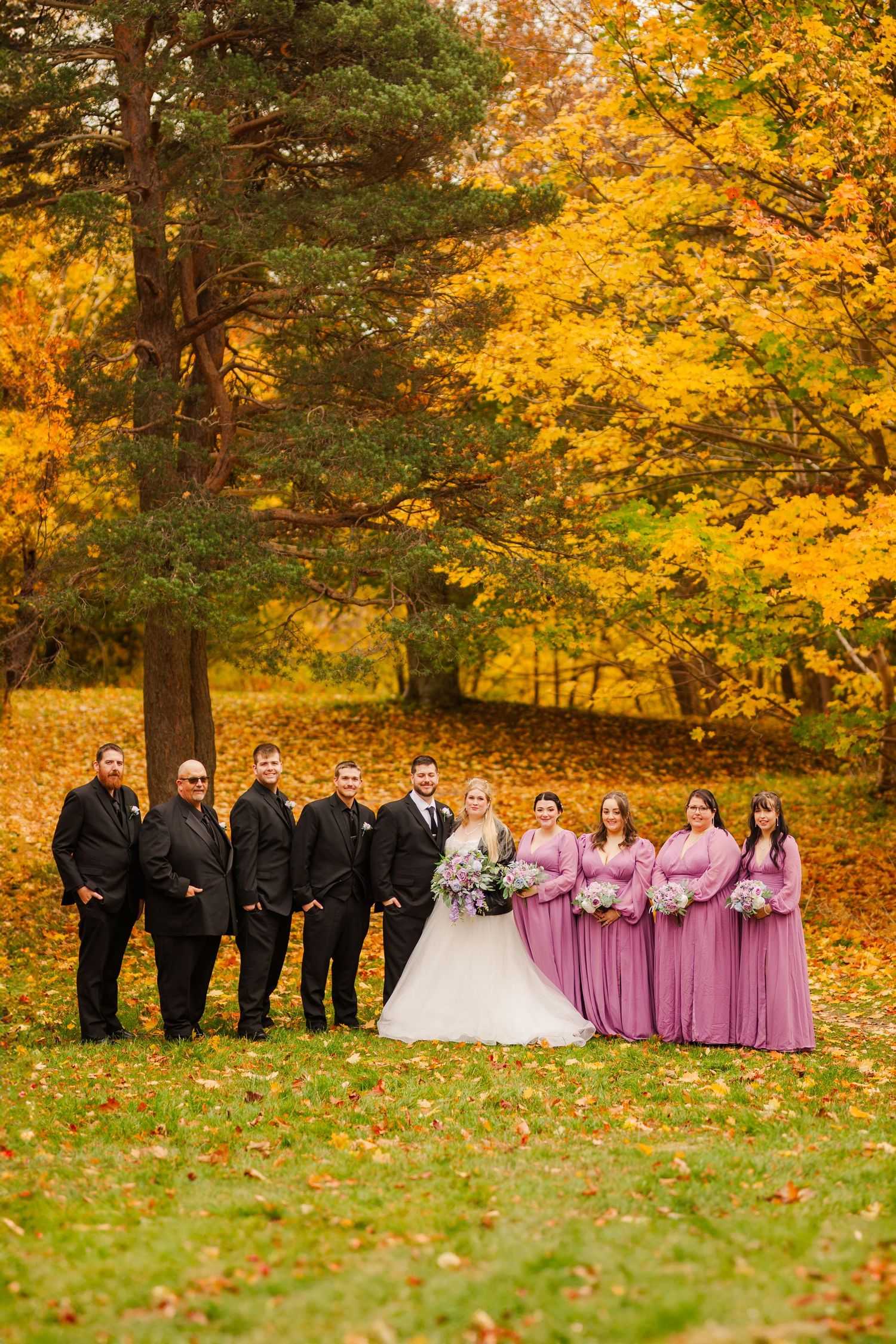 Wedding party poses in a line against vibrant autumn foliage with golden yellow leaves and evergreen trees.