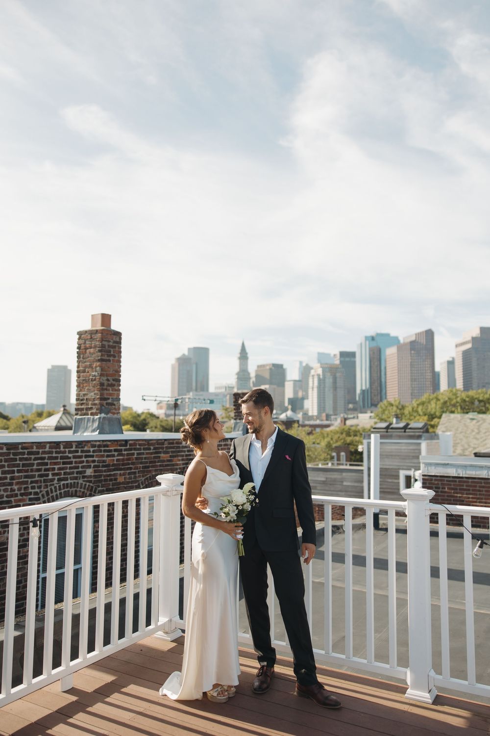 Wedding couple shares romantic moments on a rooftop deck with city skyline backdrop.