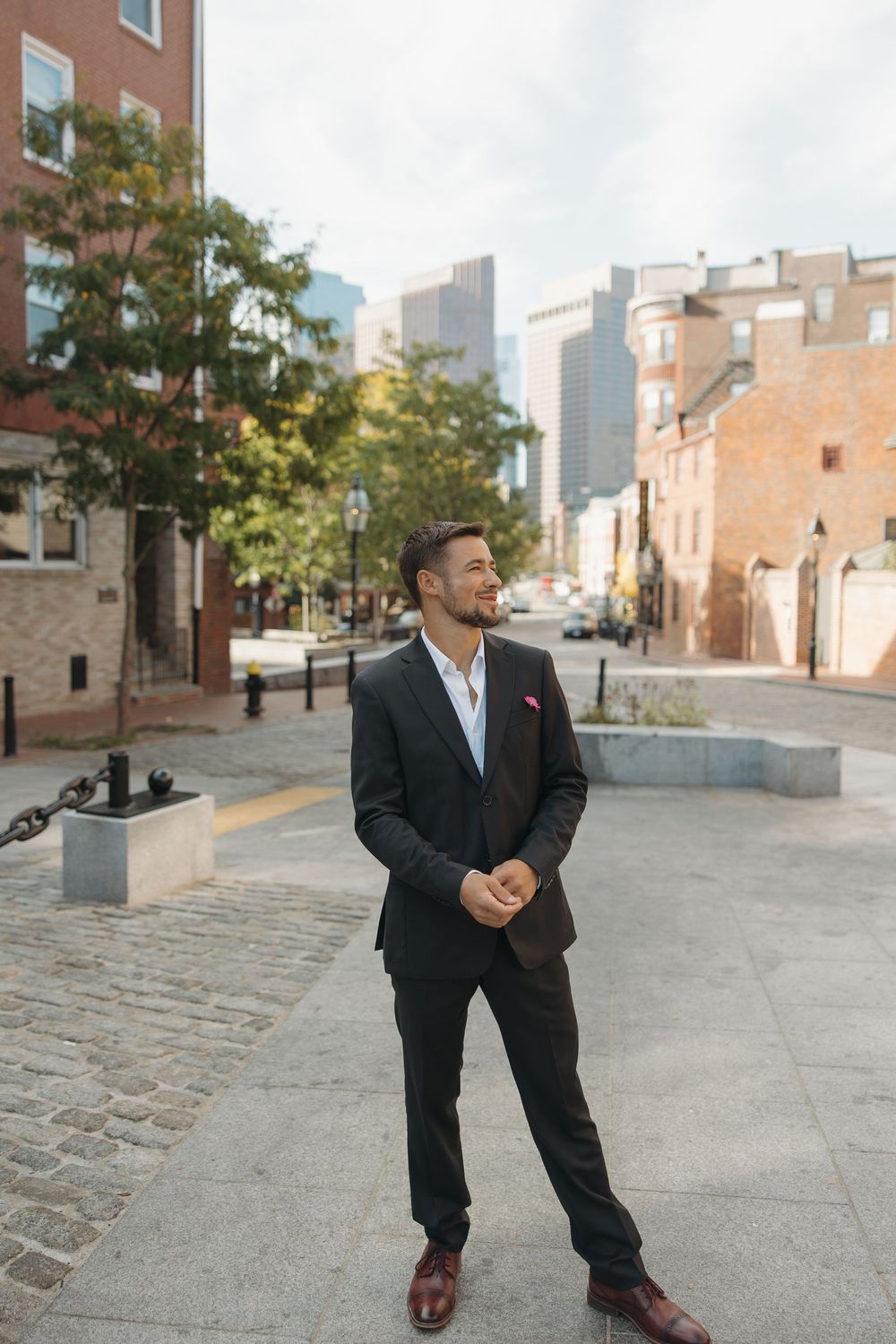 Groom in classic black suit poses in urban setting with brick buildings.