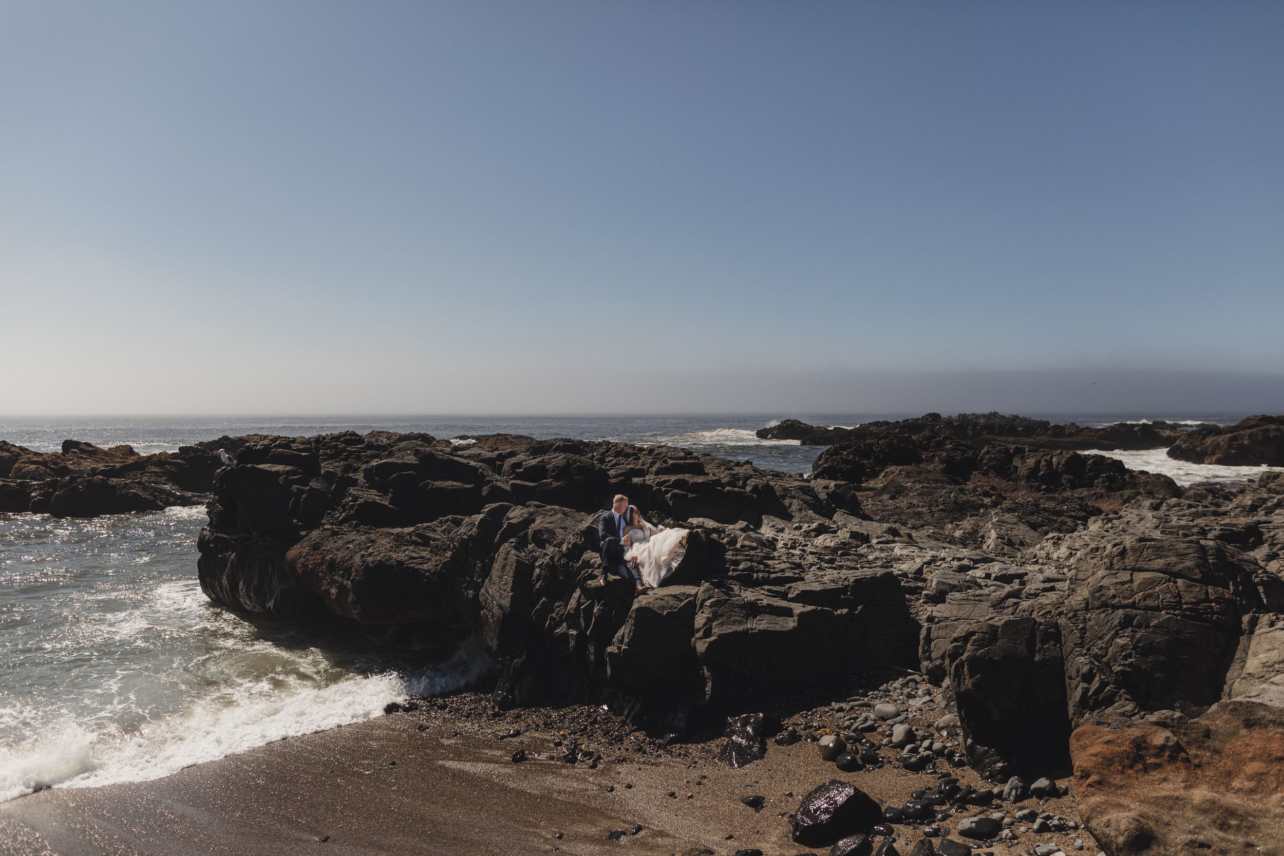 Panoramic view of rocky coastal cliffs with crashing ocean waves and distant mountains under a clear blue sky.