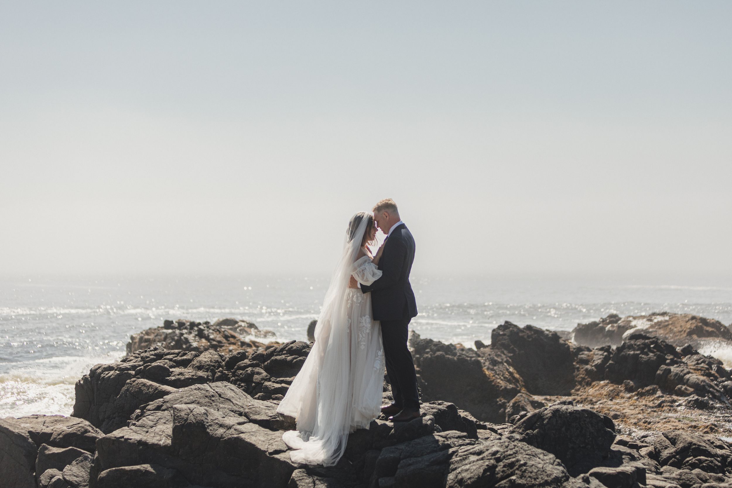 A couple in wedding attire embraces on rocky coastal cliffs overlooking a misty ocean.