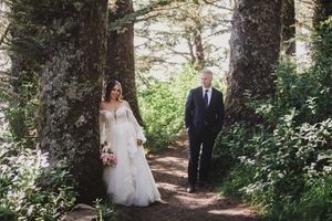 Wedding couple walks through a lush forest path surrounded by tall evergreen trees and dappled sunlight.