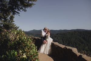 A couple stands on a scenic coastal cliff overlooking mountains and ocean on their wedding day.