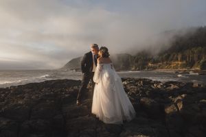 Newlyweds pose on rocky coastal cliffs with foggy ocean views in the background.